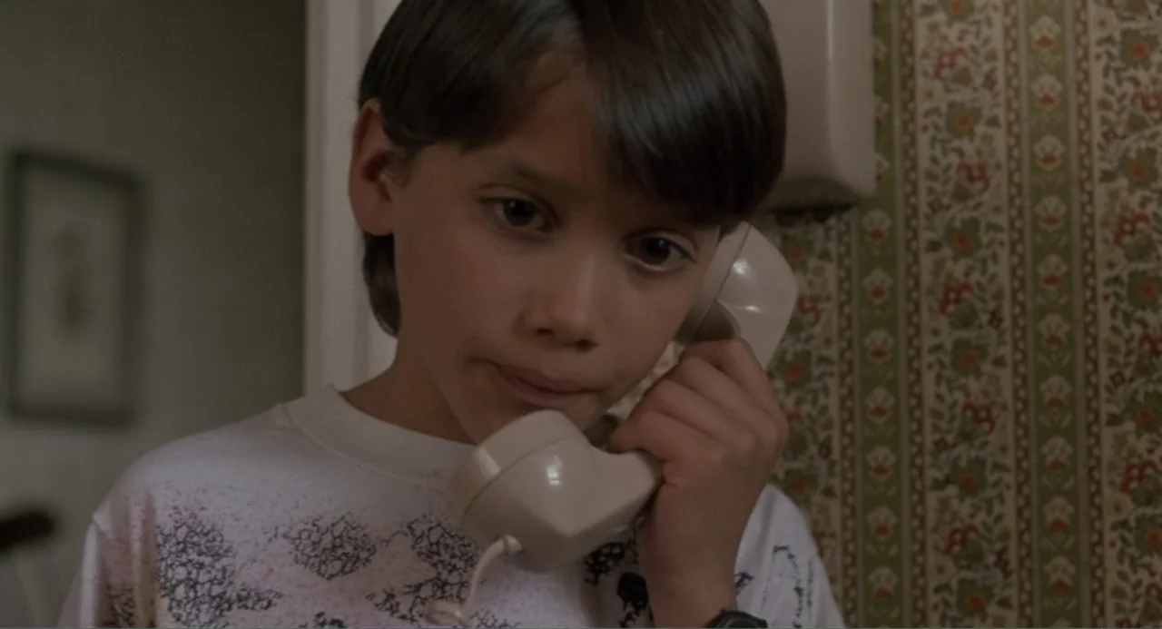 F/X2 (1991) movie still: closeup — A domestic interior features floral patterned wallpaper and a blurred background structure. A young boy holds a…