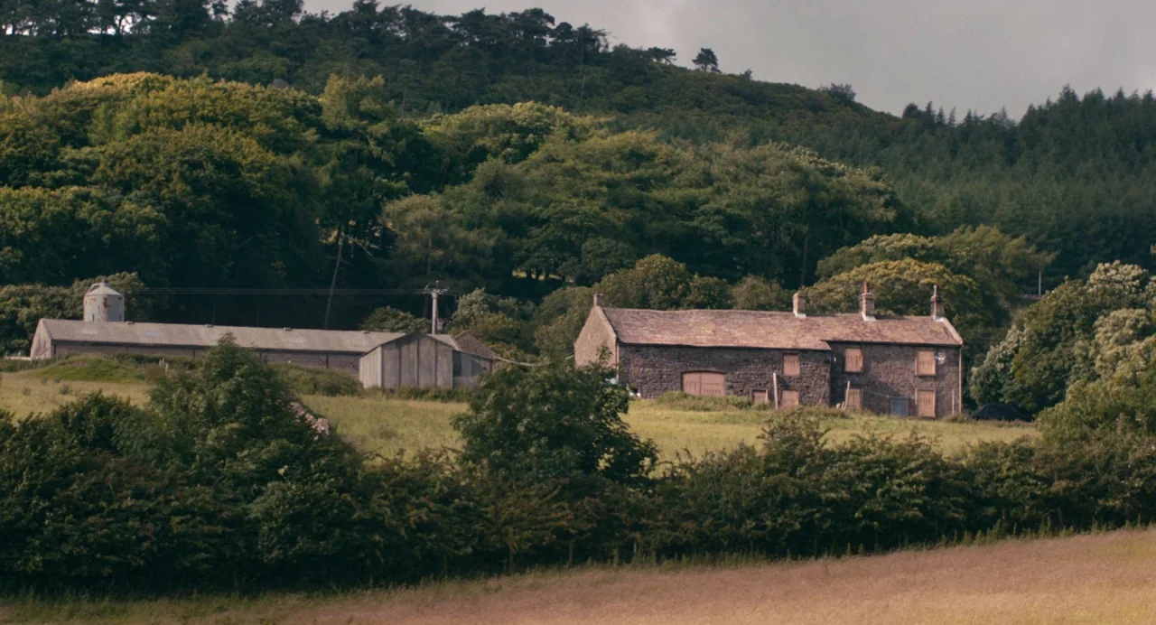 Dark River (2017) movie still: establishing — A stone farmstead sits at the base of a dense, tree-covered hillside. Multiple outbuildings and a…