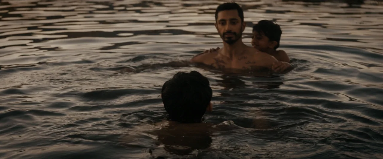 Encounter (2018) movie still: medium — An open body of water during golden hour with gentle ripples on the surface. A man with chest tattoos swims…