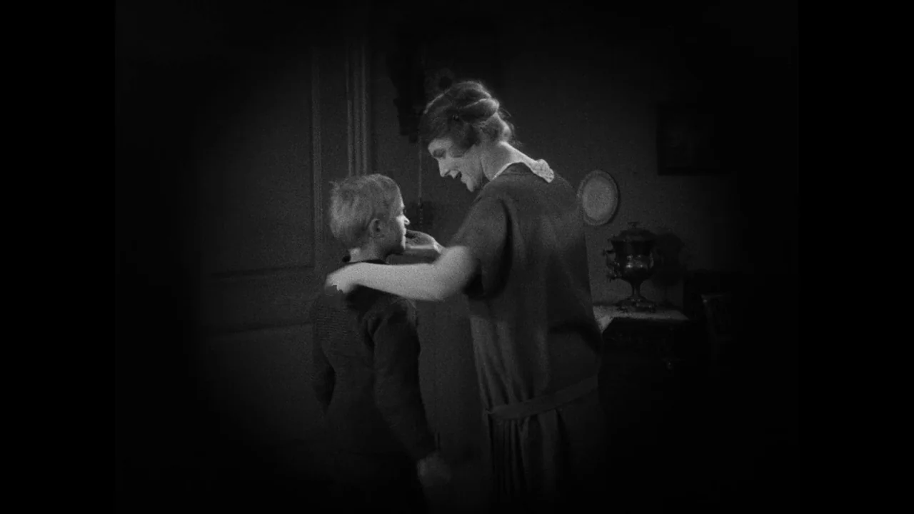 Master of the House (1925) movie still: medium — A dimly lit room contains decorative plates on the wall and a small table. A woman stands over a…