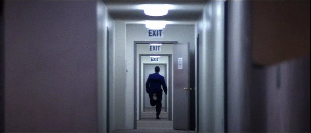Punch Drunk Love (2002) movie still: wide — A long, symmetrical series of repeating interior doorways creates a tunnel effect. A man in a dark suit…