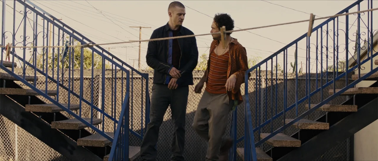 In Time (2011) movie still: fullbody — An outdoor metal staircase connects to a chain-link fenced area during the day. Two men stand together on the…
