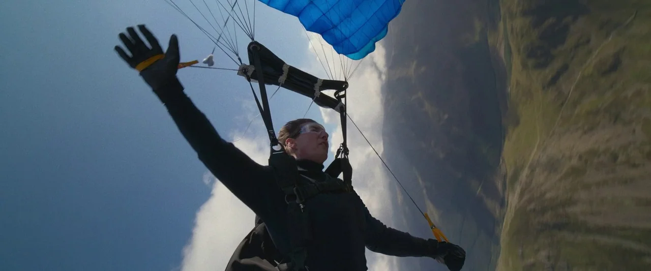 A person floats through the air using a blue parachute over mountainous landscape. The subject hangs from the harness with arms extended while wearing protective goggles.