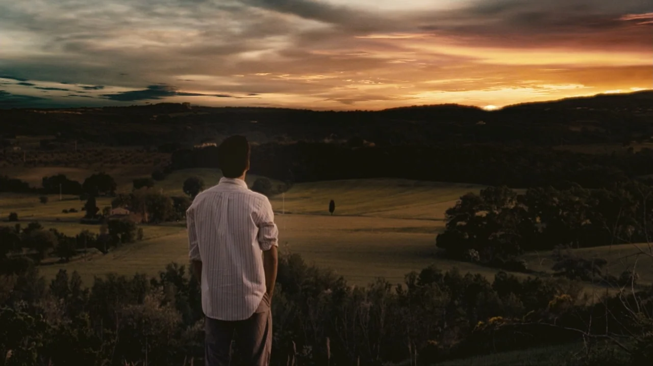 Shadows in the Sun (2005) movie still: fullbody — A rolling rural landscape with distant trees and fields under a cloudy sunset sky. A man stands…