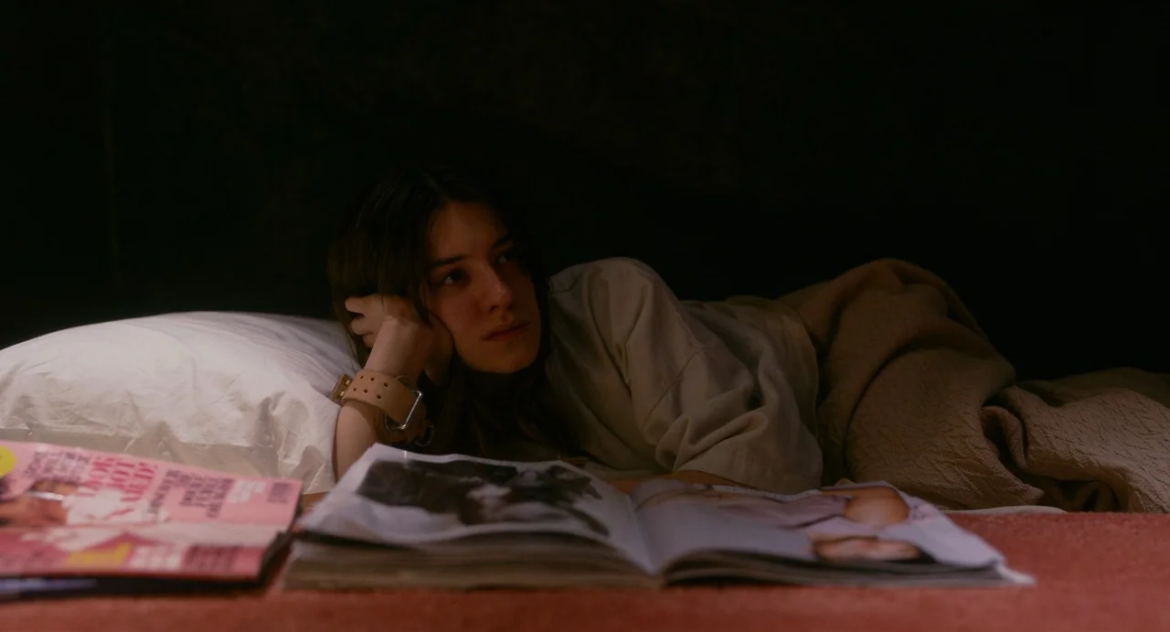 Fresh (2022) movie still: medium — A dimly lit room with a person resting on a bed covered in dark textiles. A magazine lies open in the foreground…