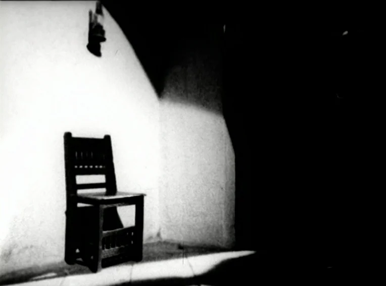 Cuadecuc, Vampir (1971) movie still: medium — A dark, spartan room features a single wooden chair placed against a white wall. A high-contrast light…