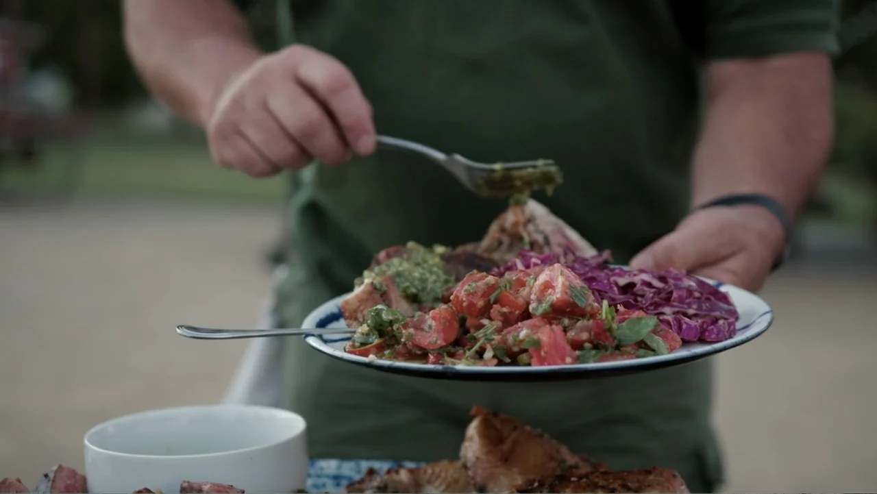 Common Ground (2000) movie still: closeup — An outdoor setting features a person holding a plate of food at a picnic table. The person uses a fork…