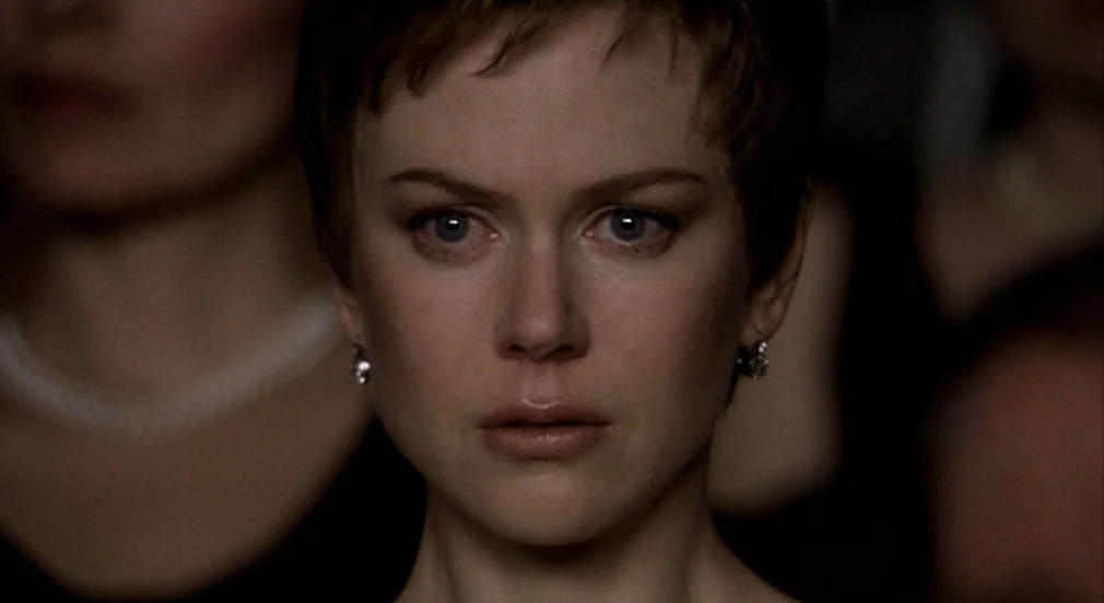 Birth (2004) movie still: closeup — A woman with short brown hair stands in a dimly lit auditorium setting. She looks forward while other blurred…