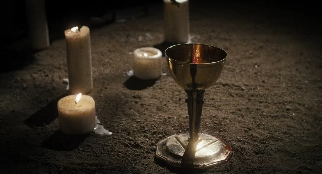 Gates of Darkness (2019) movie still: closeup — A brass chalice rests on a patch of dark, loose soil at night. Multiple lit candles surround the…
