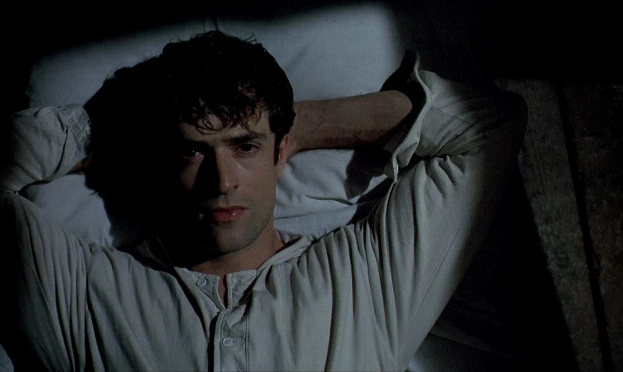 Dellamorte Dellamore (1994) movie still: closeup — A man lies on a white pillow in a dark, dimly lit environment. He rests his hands behind his head…