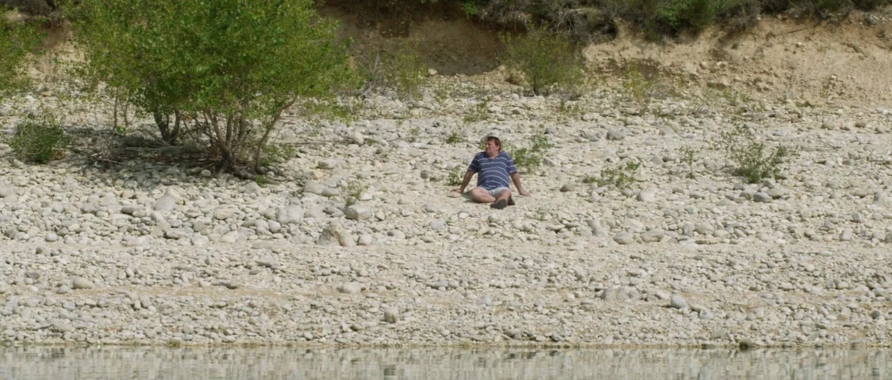 Stranger By The Lake (2013) movie still: wide — A dry, rocky riverbank with scattered vegetation and a high, sandy embankment behind. A man in a…