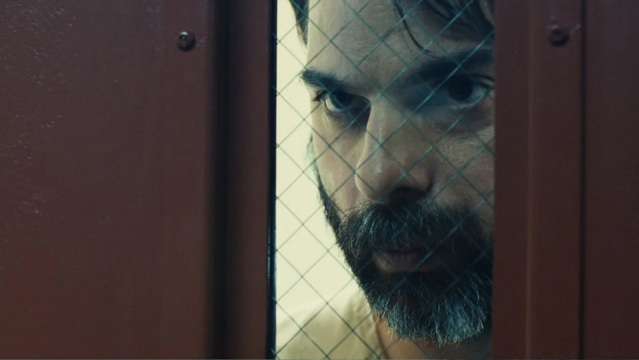 Camp X-Ray (2014) movie still: closeup — A visitation room featuring a thick glass partition with integrated wire mesh. A bearded man looks through…