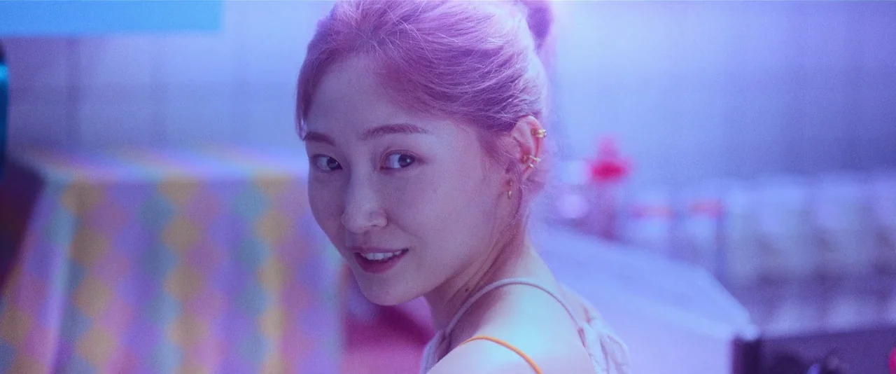 Ballerina (2023) movie still: closeup — A room with violet artificial lighting contains a patterned tablecloth in the background. A young woman with…