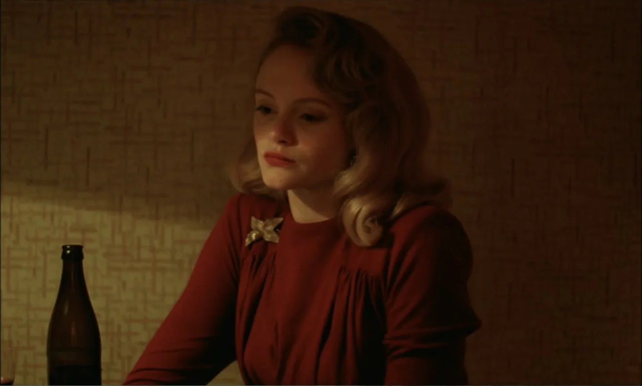 The Brutalist (2024) movie still: medium — A dimly lit room with textured beige wallpaper and a dark table. A blonde woman in a red dress sits…