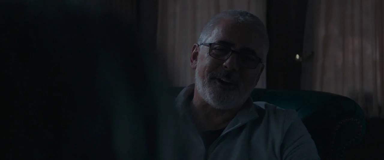 Pig (2021) movie still: medium — A dimly lit room with draped curtains visible in the background. An older man with glasses sits and speaks toward…