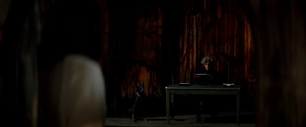 In The Mouth of Madness (1994) movie still: wide — A dark office features a metal desk and a backdrop with textured reddish-orange panels. A man…
