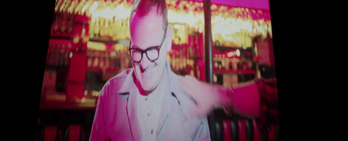 Cheap Thrills (2013) movie still: medium — A dimly lit diner booth at night features intense pink neon lighting behind subjects. A man wearing…