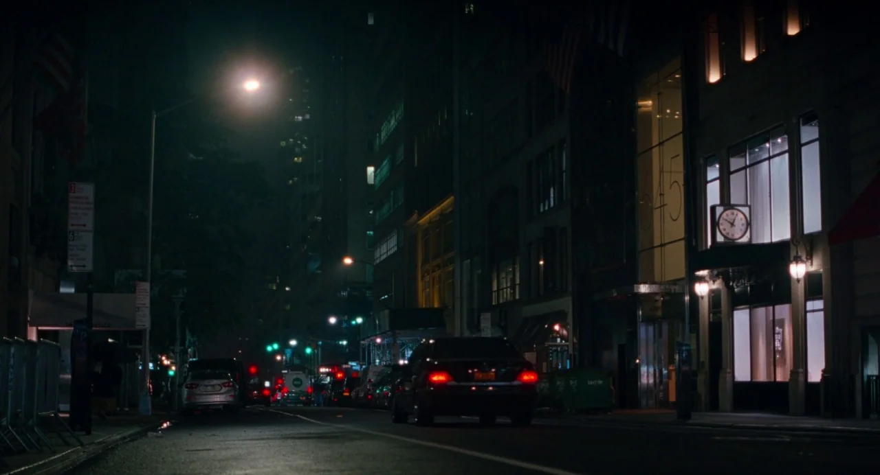 On The Rocks (2020) movie still: wide — A dark, quiet urban street at night is lined with tall commercial buildings. A black car drives away from…
