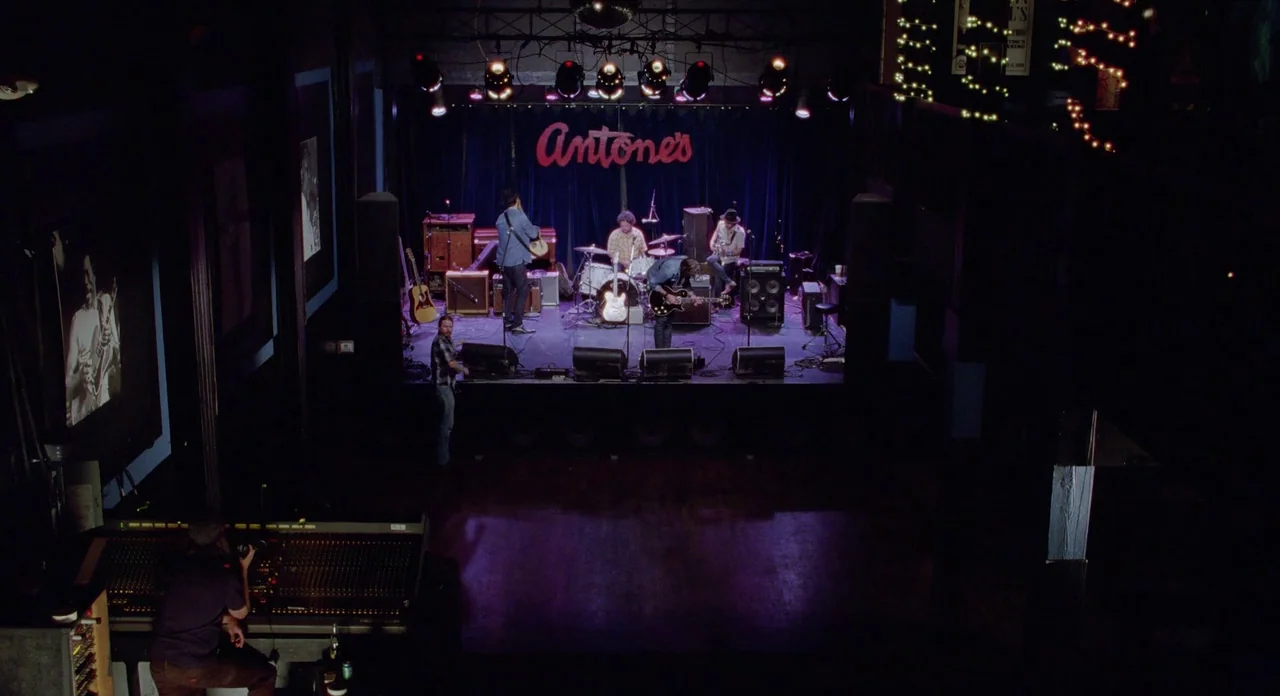 Boyhood (2014) movie still: wide — A dark, cavernous music club features a raised stage with a large neon sign. Four musicians play instruments on…