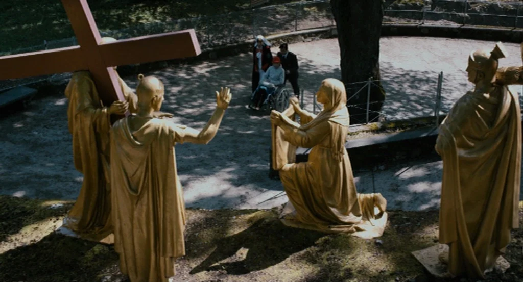 Lourdes movie still: wide — A gravel pathway features several large, gold-colored statues depicting biblical figures. A group of three people walks…