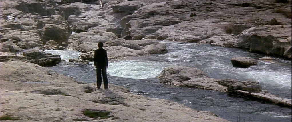 Love Unto Death (1984) movie still: wide — A rugged river gorge features light gray limestone rock formations surrounding a flowing stream. A lone…