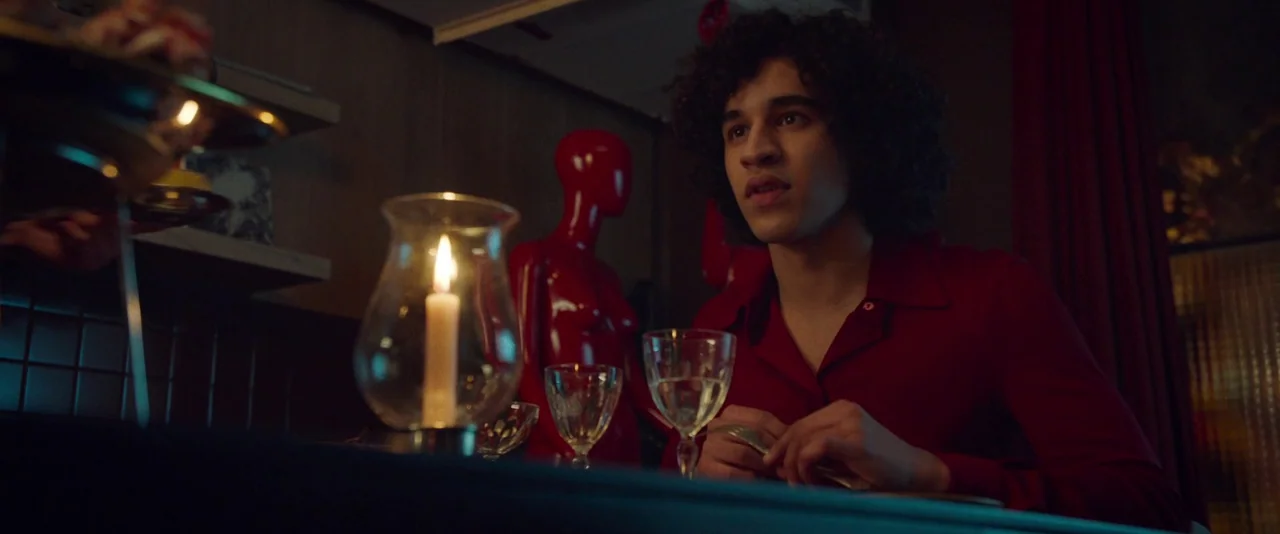 Peter Von Kant (2022) movie still: medium — A dimly lit room features a table set with a burning candle and glassware. A young man with dark curly…