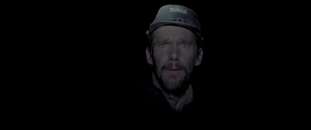 In A Valley of Violence (2016) movie still: closeup — A man with a goatee wearing a kepi hat stares out from black darkness. The close framing…