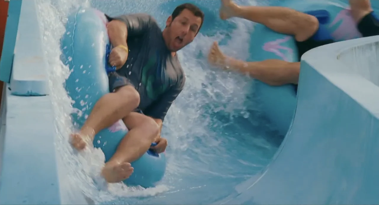 Grown Ups (2010) movie still: medium — A light blue fiberglass water slide has rushing water moving over its surface. An adult man rides an…