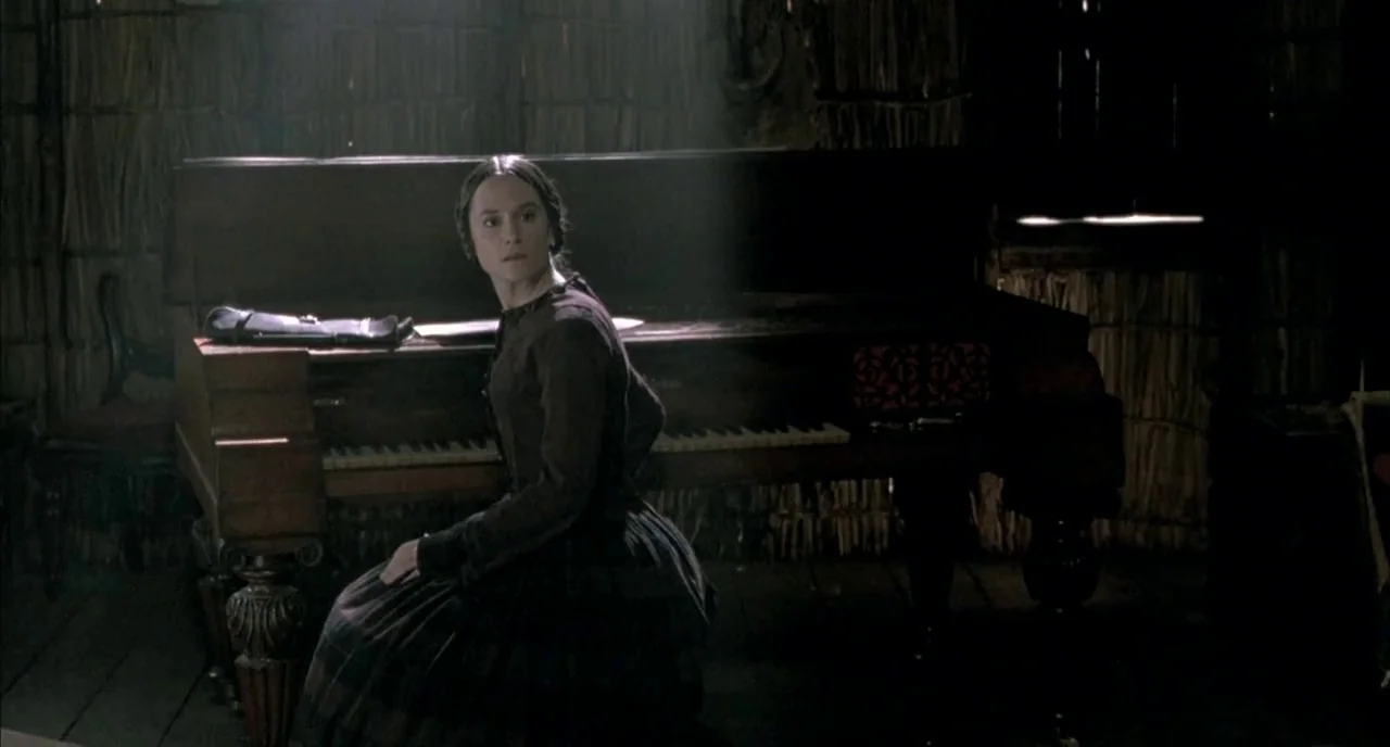 The Piano (1993) movie still: medium — A dark, dimly lit room features walls with textured, vertical paneling. A woman in a dark period dress sits…