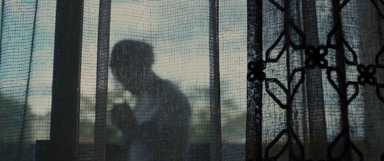 Sicario (2015) movie still: medium — A silhouette of a person stands behind a translucent, patterned curtain by a window. The person is captured in…