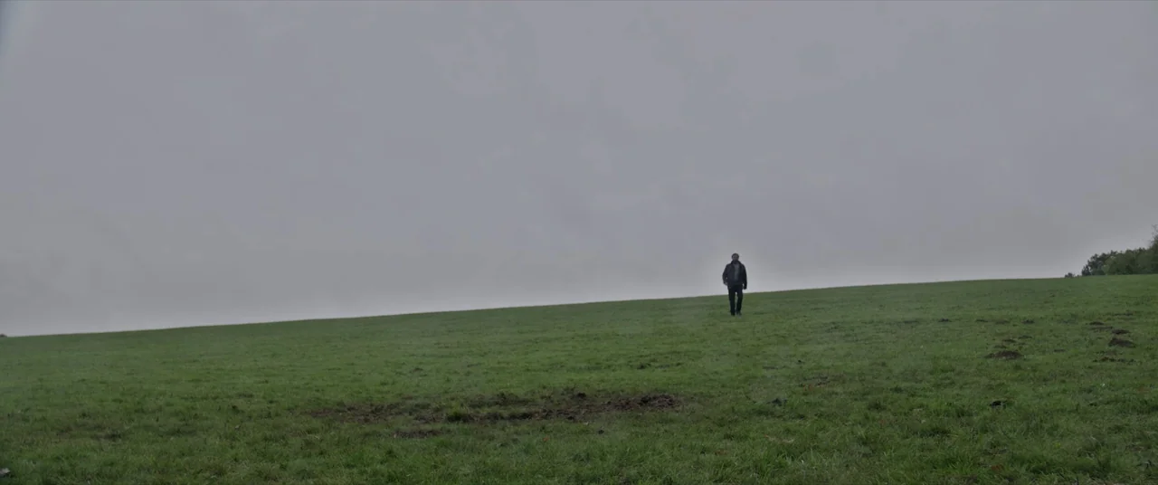 Bull (2021) movie still: wide — A vast, empty green hill stretches toward an overcast, grey sky. A single man walks slowly across the incline of the…