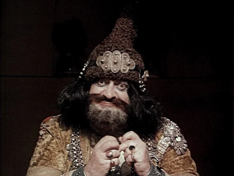 Ashik Kerib (1988) movie still: closeup — A man wearing an ornate hat and traditional vest stands in a dark space. He stares forward while holding a…