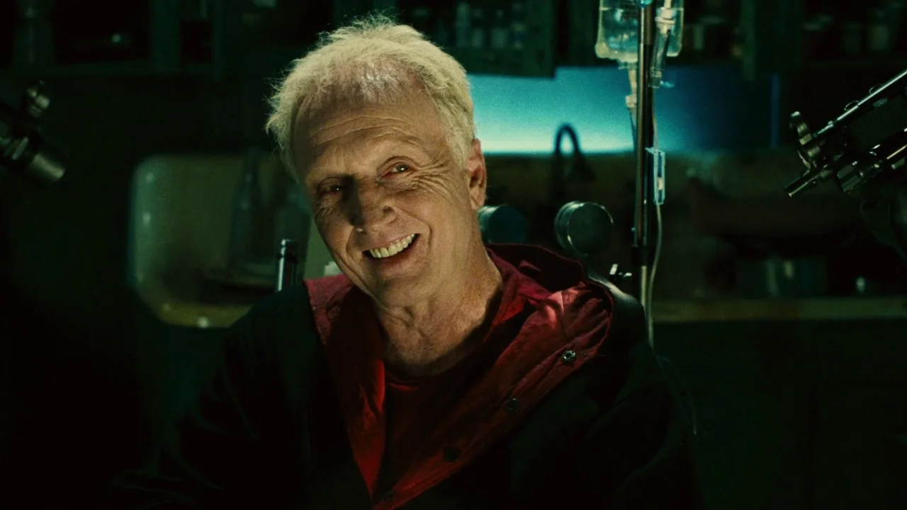 Saw II movie still: closeup — A dimly lit room with medical equipment and an intravenous drip stand visible. An older man sits in a chair looking…
