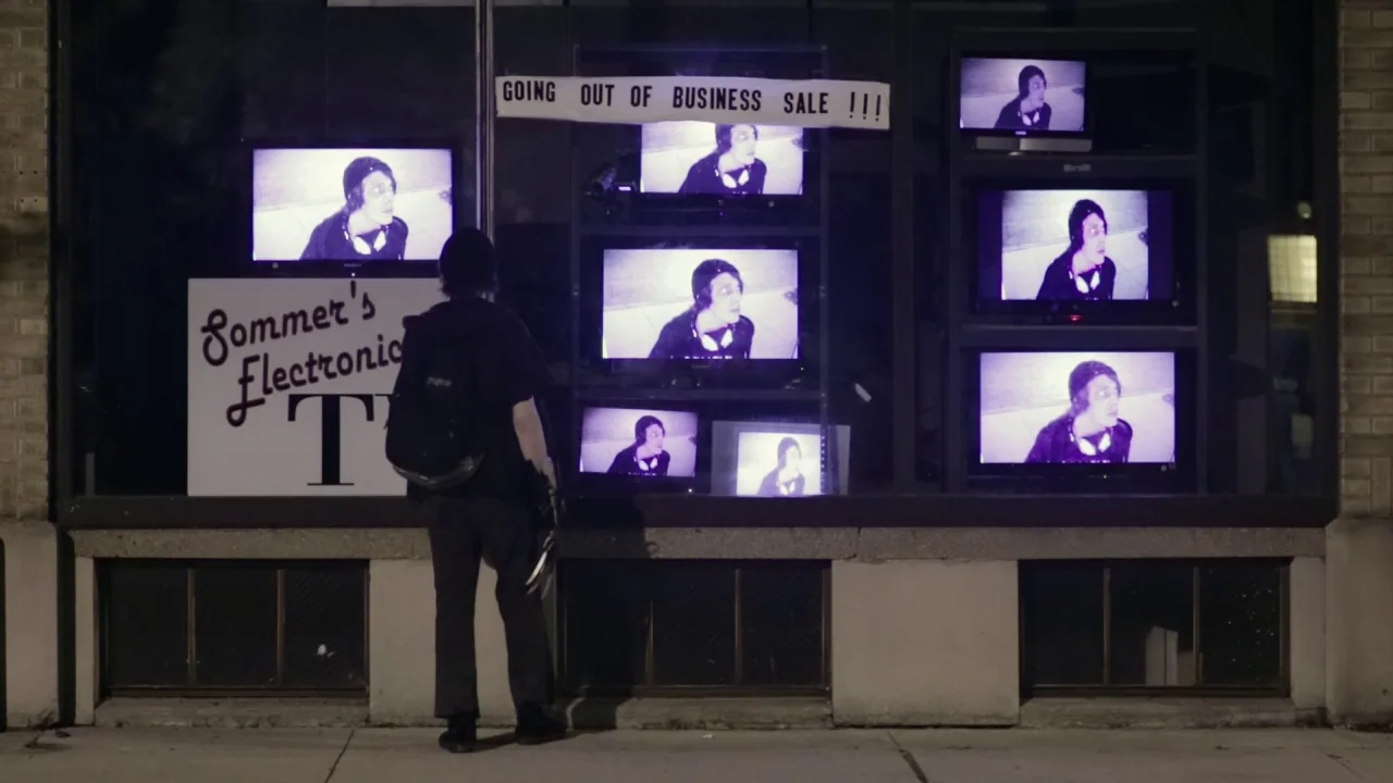 Buzzard (2014) movie still: fullbody — A dark city storefront features a window display with numerous televisions playing identical footage. The…