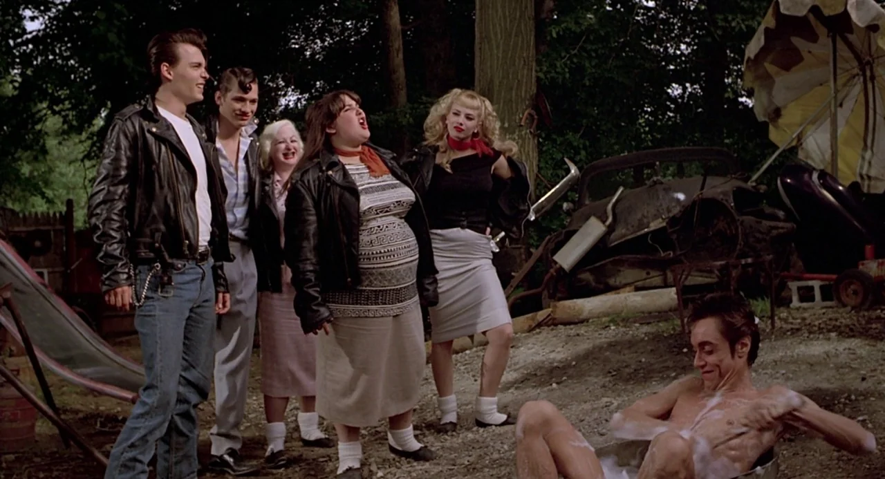 Cry Baby (1990) movie still: wide — An overgrown backyard contains a rusted vehicle frame, old wood, and scattered garden debris. A group of…
