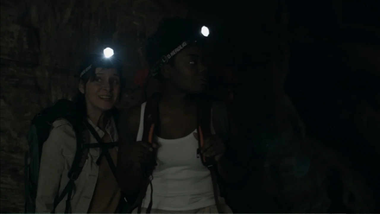 The Grotto (2022) movie still: medium — Two women stand in a dark, narrow rocky cavern holding backpacks and headlamps. They look around the dark…
