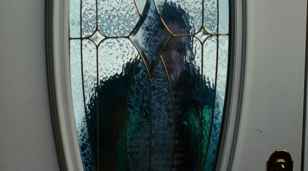 Juno (2007) movie still: closeup — A view through a textured, leaded glass panel in an oval front door. A person stands on the other side, visible…