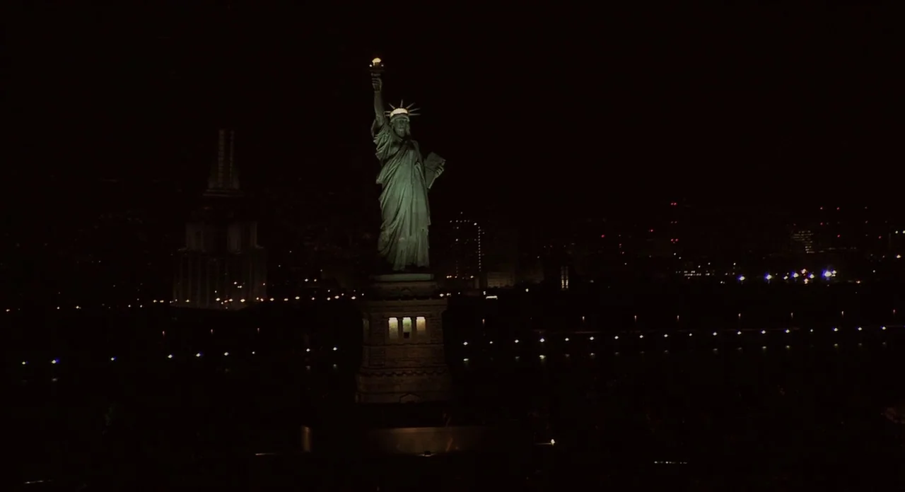 Populaire (2012) movie still: wide — The Statue of Liberty stands centered in a dark harbor at night. Distant city lights glow faintly along the…