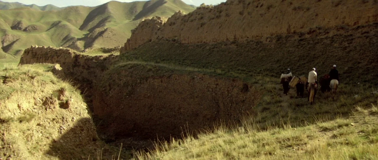 Horse Thief movie still: wide — A group of travelers on horseback traverses a narrow earthen trail between steep hills. The arid landscape features…
