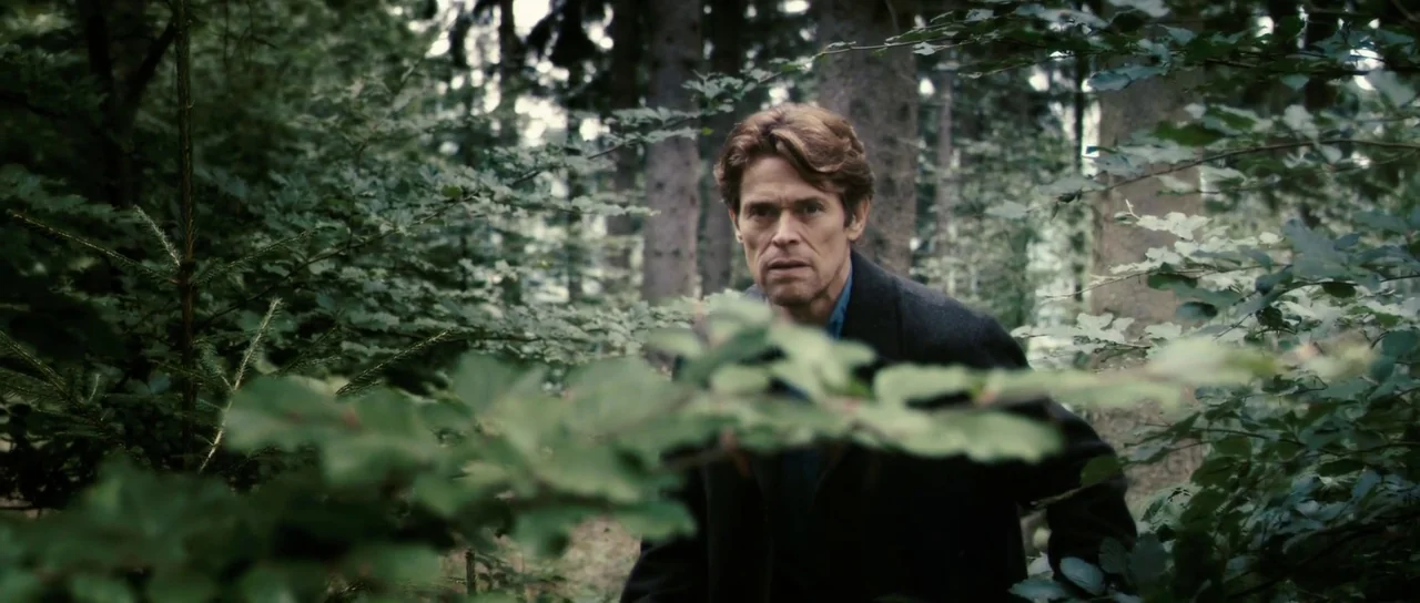 Antichrist (2009) movie still: medium — A dense patch of temperate woodland features heavy green foliage and tall pine trees. A mature man in a dark…