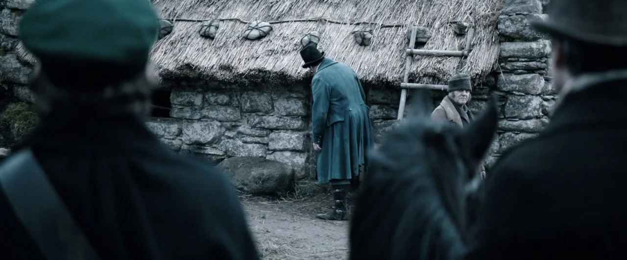 Black ’47 (2018) movie still: wide — A rustic stone dwelling with a thatched roof sits against a rocky terrain background. A man in a top hat…
