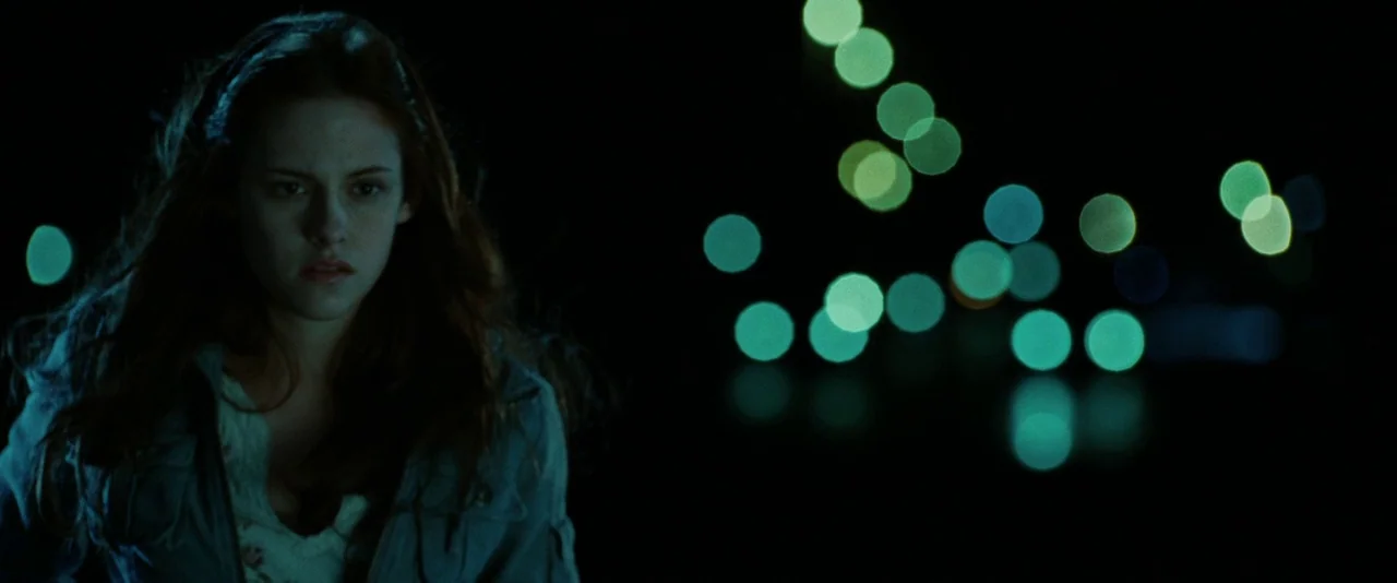 Twilight (2008) movie still: medium — A dark outdoor space at night serves as the backdrop for a lone young woman. Out-of-focus street lights create…
