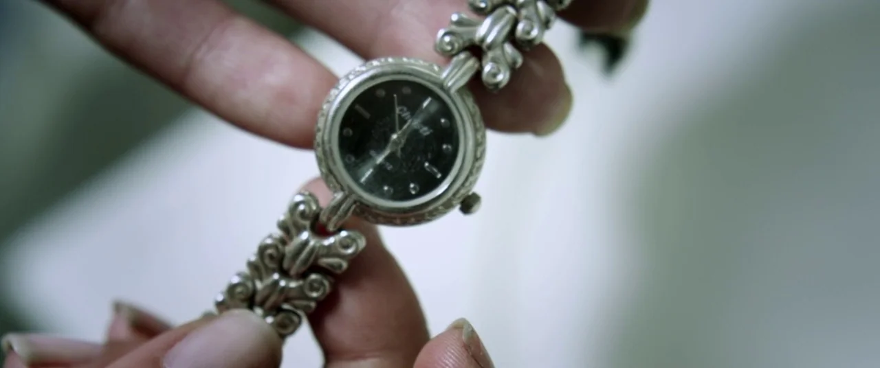Here Comes The Devil (2012) movie still: closeup — A silver watch with a dark face is held between two sets of fingers. The background is a clean,…