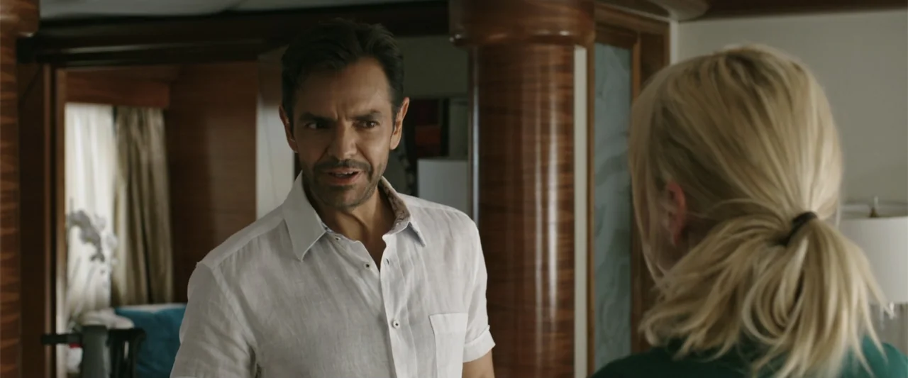 Overboard (2018) movie still: medium — A luxury interior cabin with polished dark wood paneling and beige fabric curtains. A man faces a woman who…