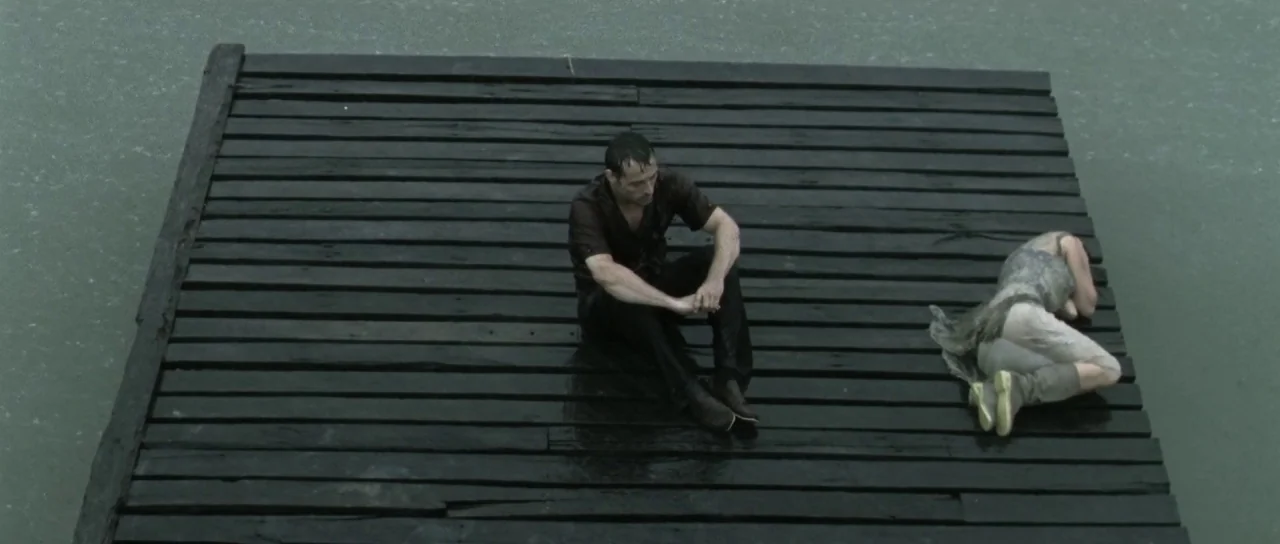 Vinyan (2008) movie still: wide — A dark wooden pier floats on the surface of calm, grayish-green water. A man sits upright near a woman lying…