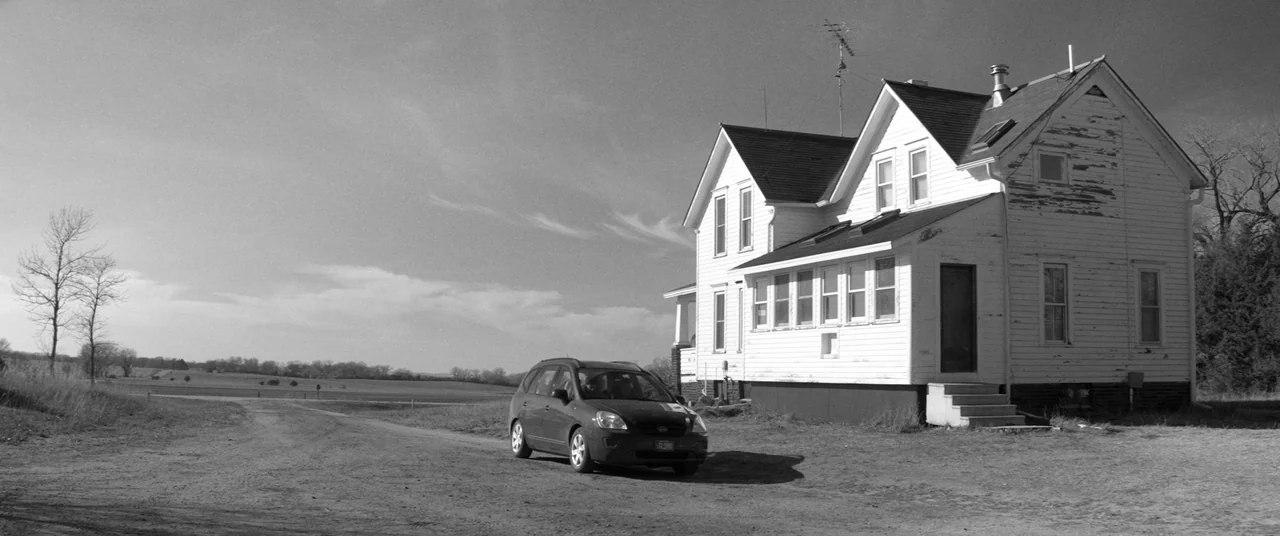 Nebraska (2013) movie still: wide — A weathered white farmhouse with peeling paint stands in a vast, open field. A dark minivan is parked on the…