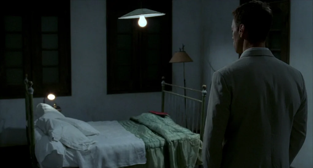 Anatomy of Hell (2004) movie still: medium — A dimly lit room contains an iron-framed bed with white linens and green blankets. A man in a light…