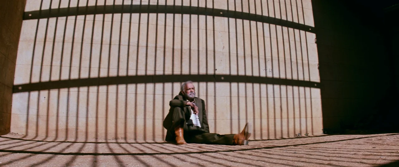 Razorback (1984) movie still: wide — A light-colored stone wall displays the sharp, grid-like shadows of unseen metal bars. An elderly man sits…