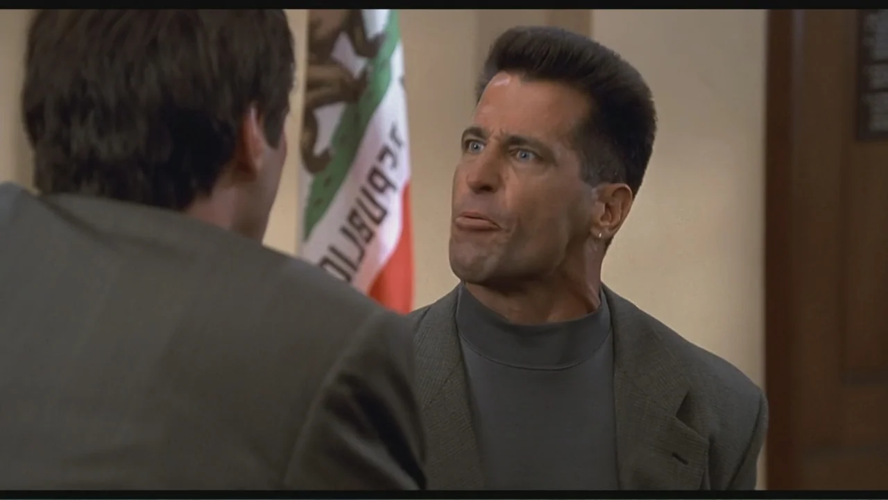 Liar Liar (1997) movie still: medium — An interior space features a California state flag displayed on a plain wall. A man in a grey mock-neck shirt…