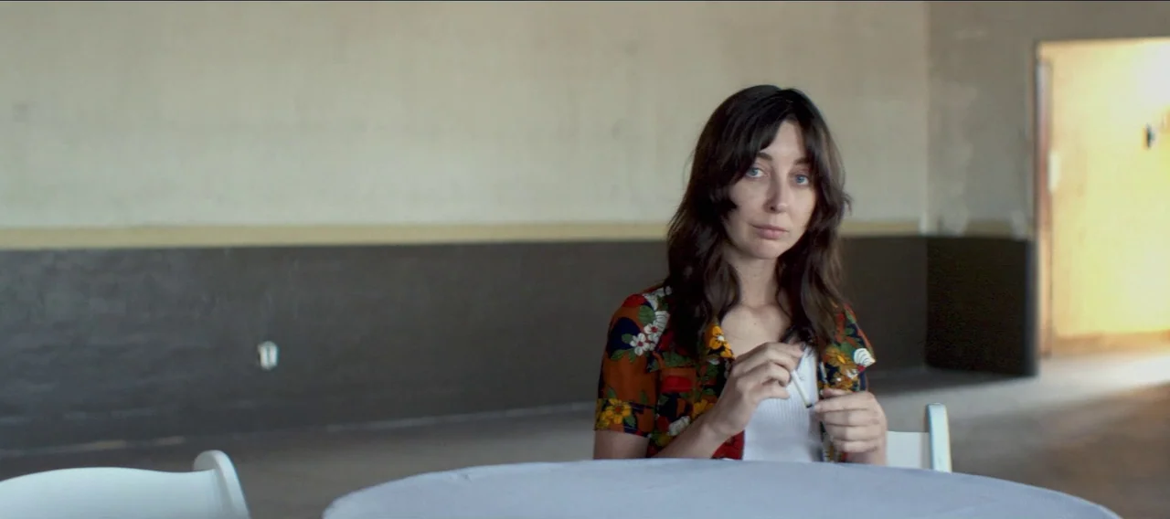 Fuzzy Head (2023) movie still: medium — A spacious room with plain walls painted half-white and half-brown. A woman sits at a round table, holding a…